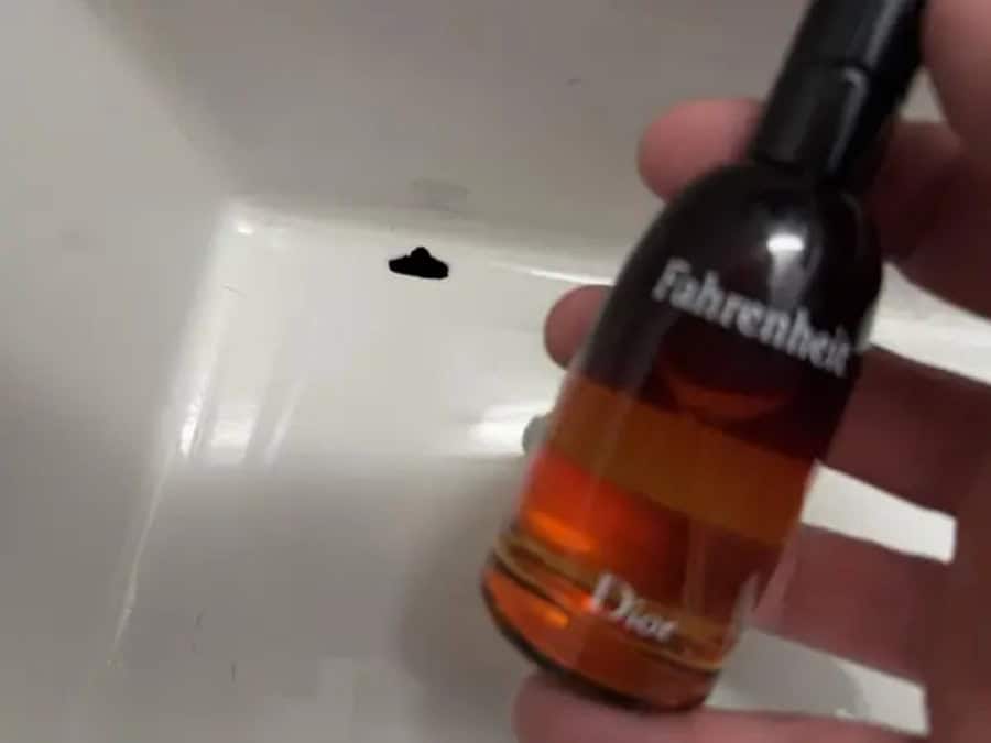 A hand holding a small bottle of Fahrenheit Dior perfume near a white surface with a black spot.