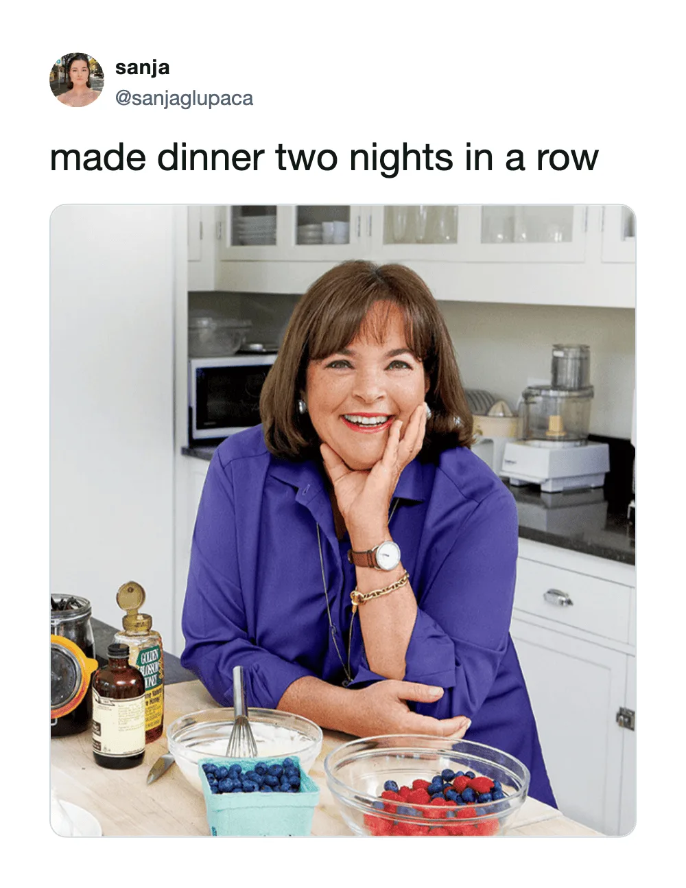 Woman in blue shirt smiling in kitchen with bowls of berries and ingredients.