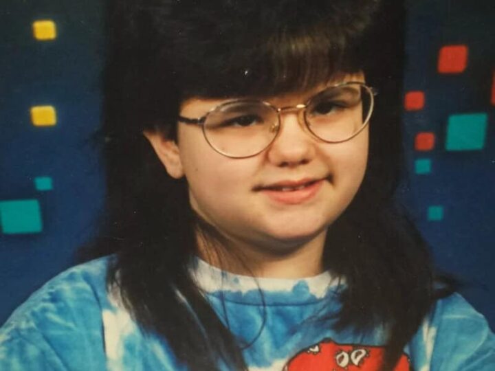 Child with big hair, glasses, and a colorful shirt, smiling in front of a patterned background.