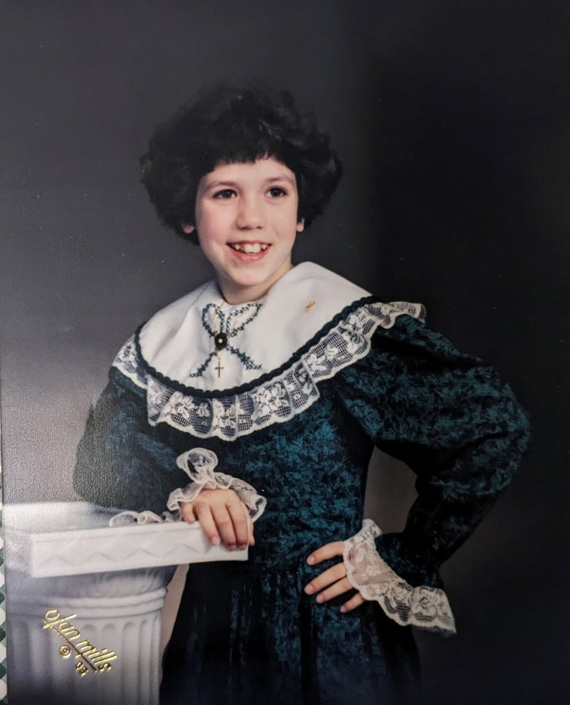 Young girl in vintage dress with lace collar and cuffs, smiling, with dark curly hair.