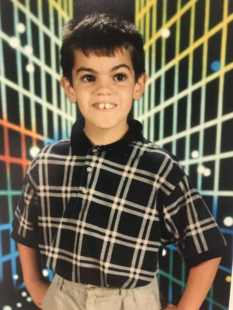 Young boy with dark hair and a checkered shirt smiling in front of a colorful grid background.