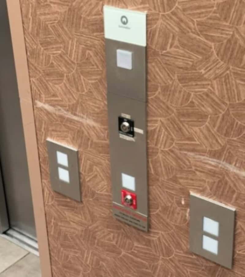 Elevator control panel with misplaced buttons and switches on a wood-patterned wall.