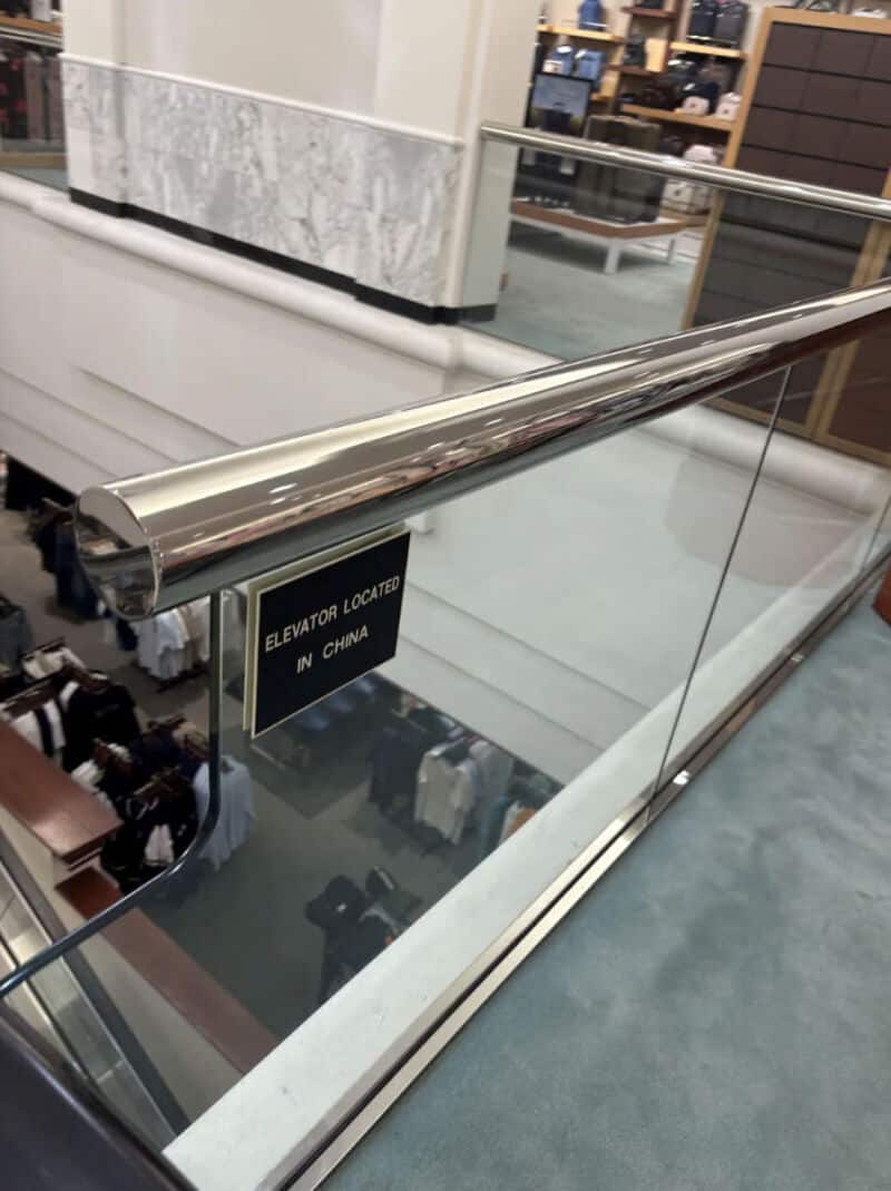Shiny metal handrail on glass balcony in a store, with a sign saying 'Elevator located on China'.