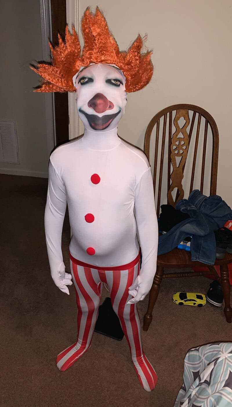 Child in clown costume with orange hair, white face paint, red nose, and striped pants.