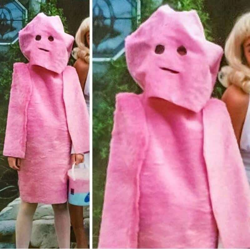 Person in pink costume with a large head and simple face, holding a bucket.