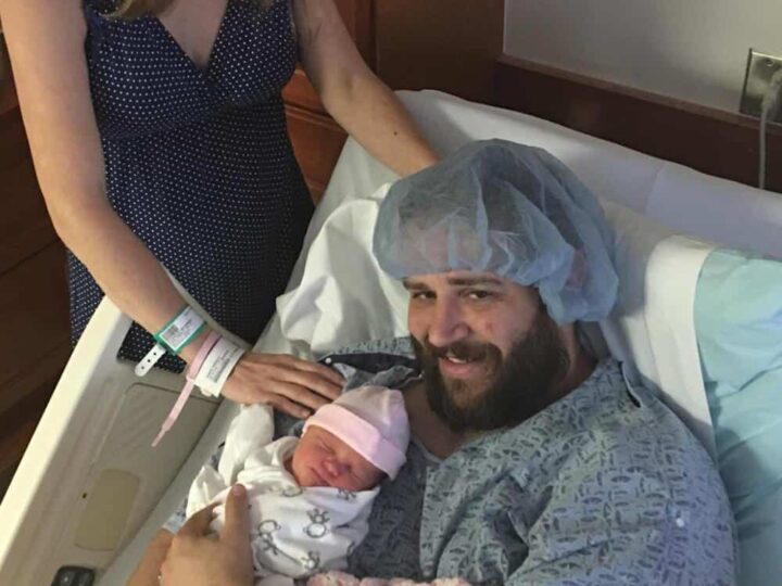 Happy woman with hospital wristbands, man in hospital gown with cap, holding newborn baby.
