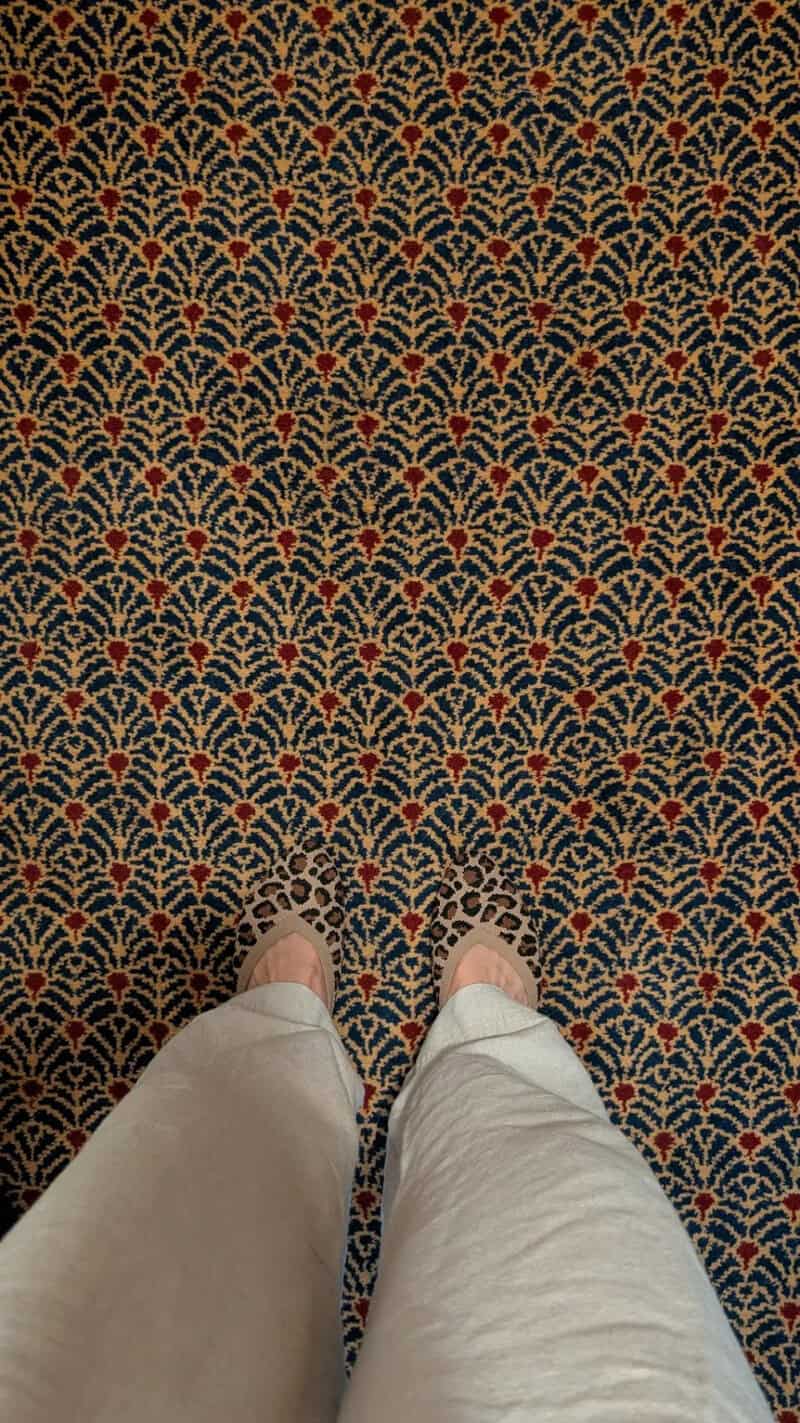 View of patterned carpet with person's feet in beige pants and white shoes.