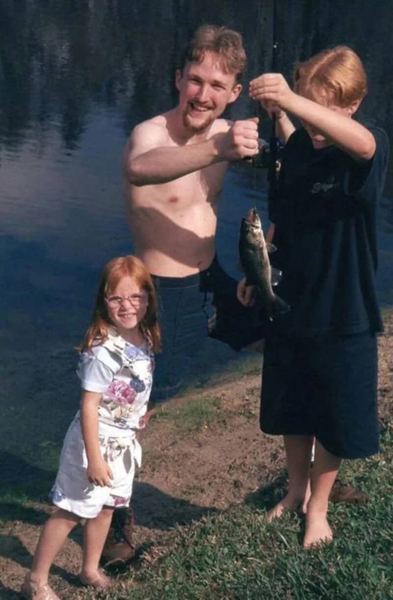 Three people by a lake, one holding a fish, with a girl smiling nearby.