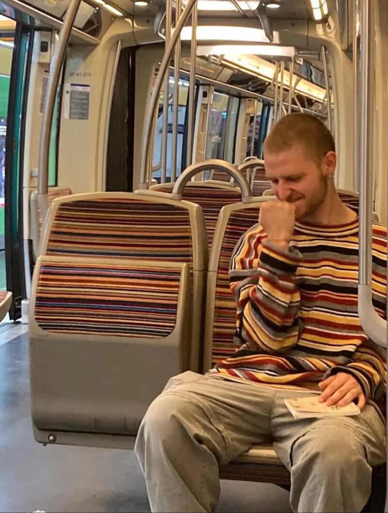 Young man with a beard in a striped sweater sitting on a bus, smiling and looking down.