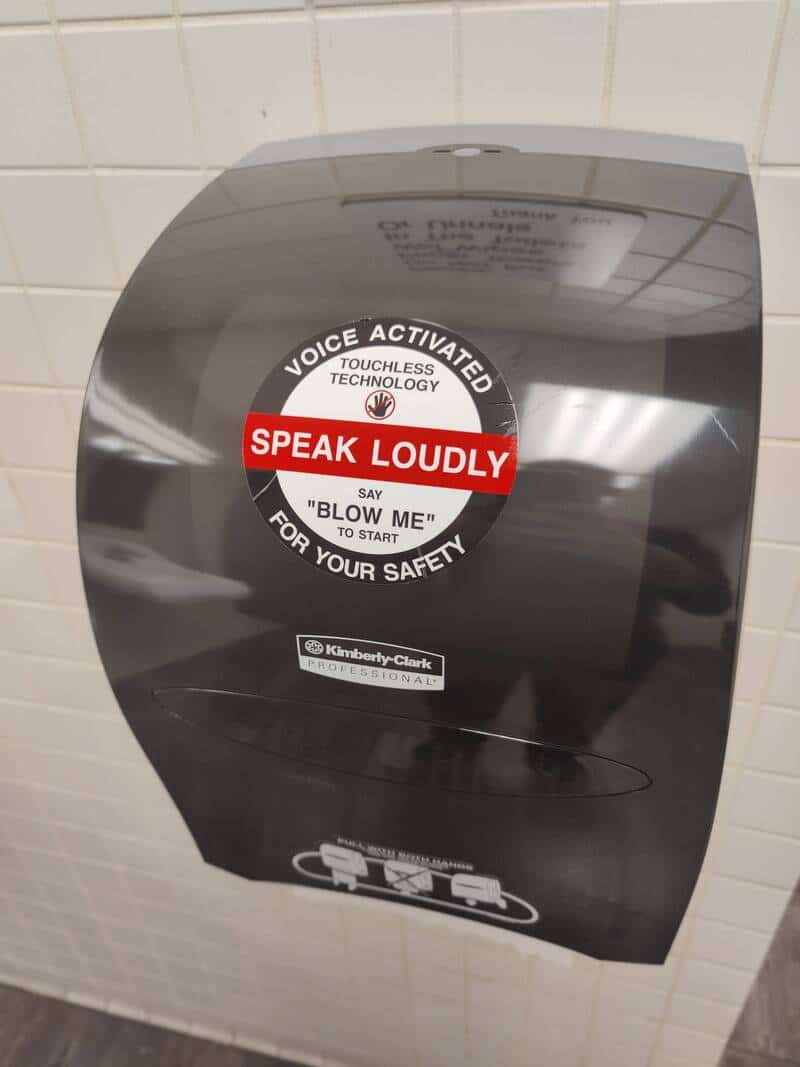 Automatic hand dryer with a sign saying 'Speak Loudly' and 'Voice Activated' in a bathroom.