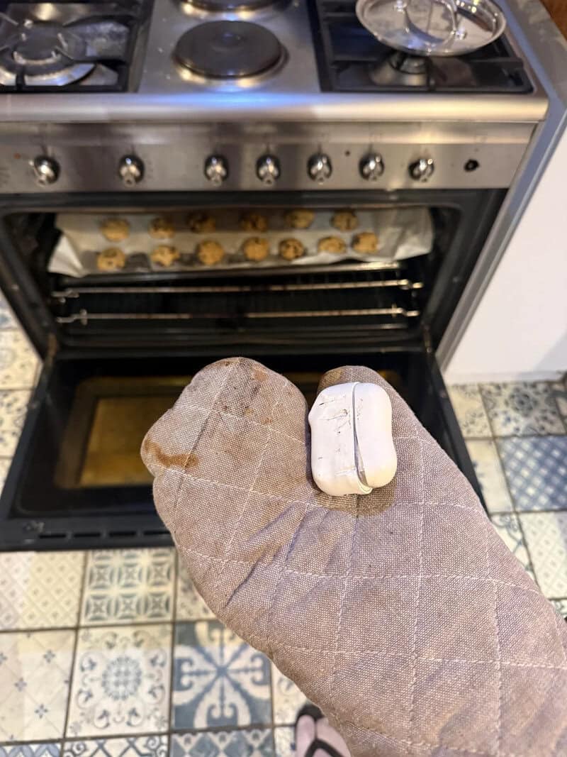 Person holding a small, white, rectangular object in front of an oven with cookies inside.