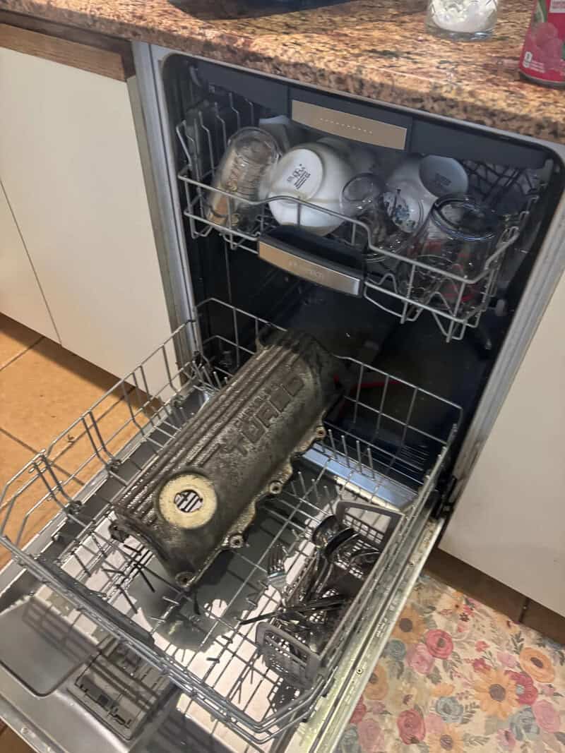 A dishwasher filled with dirty dishes, including glasses, mugs, and a large black object.