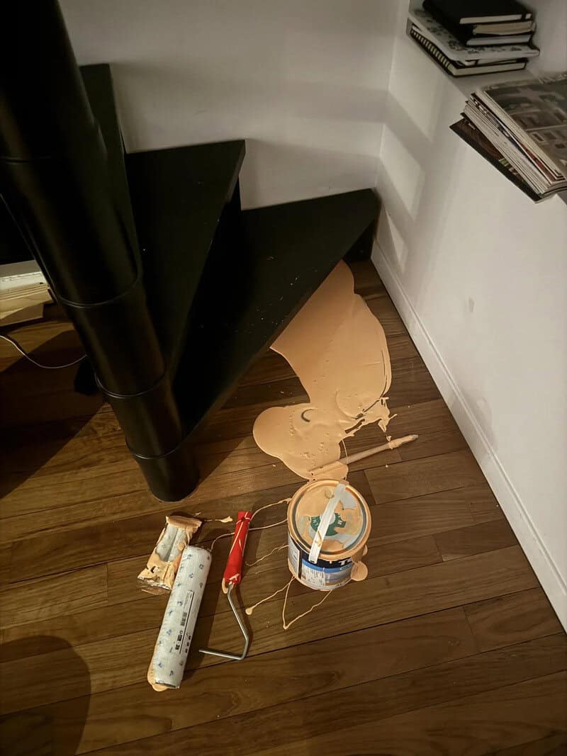 Paint spilled on wooden floor with roller and bucket nearby under black shelf.