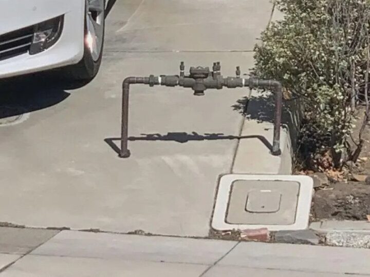 Gas meter and pipe obstructing sidewalk, near a white car and bushes.