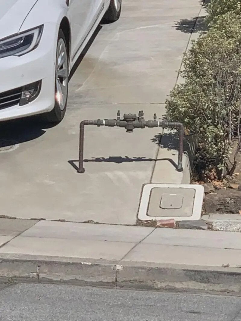 Gas meter and pipe obstructing sidewalk, near a white car and bushes.