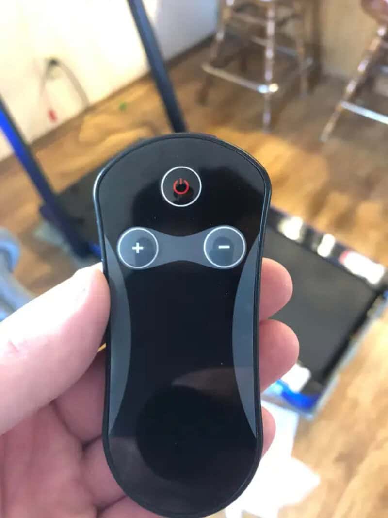Remote control with power, plus, and minus buttons in a room with wooden floor and chairs.