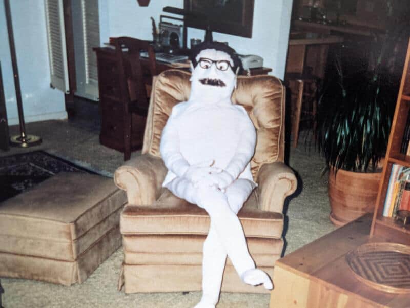 A person in a white costume with glasses and a mustache sitting on a beige armchair.