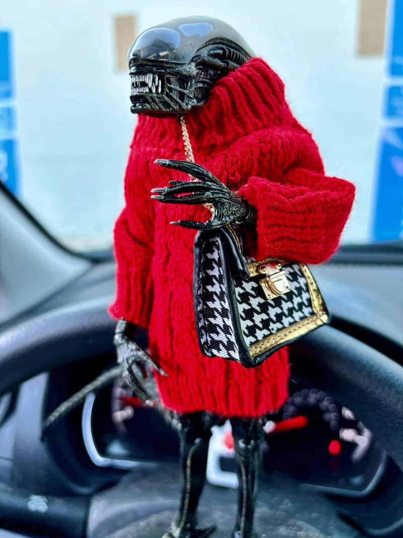Alien doll in a red sweater with a handbag inside a car.