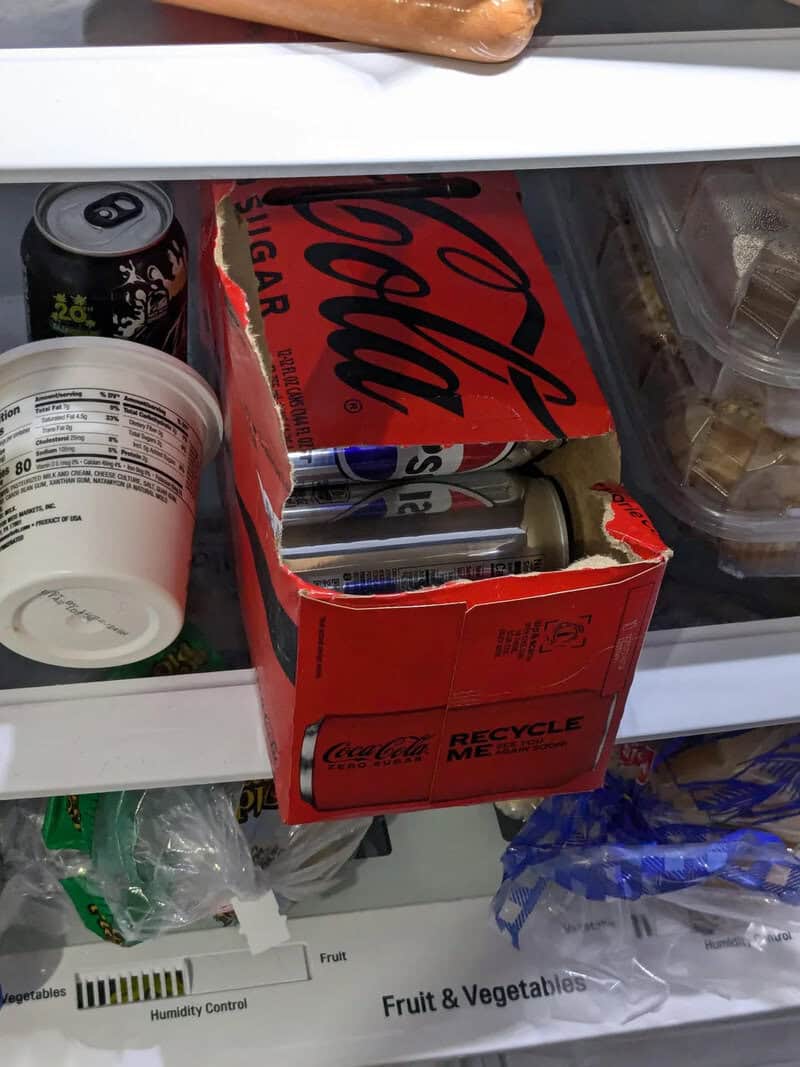Open fridge with Coca-Cola box, soda cans, and food containers inside.
