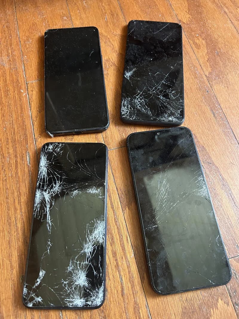 Four smartphones with cracked screens on a wooden surface.